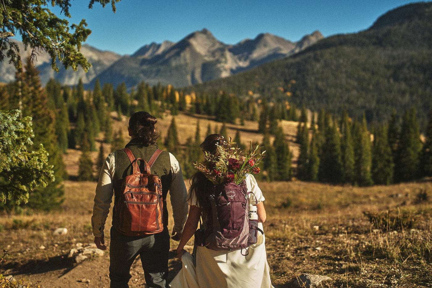 bride and groom hiking to the wedding ceremony with a backpack of flowers 