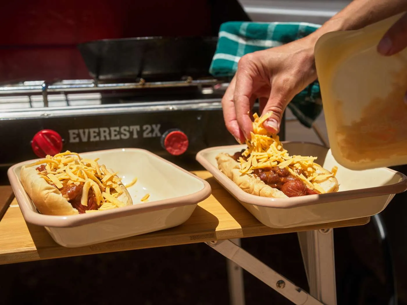 topping two chili dogs with shredded cheddar cheese after cooking over a camp stove
